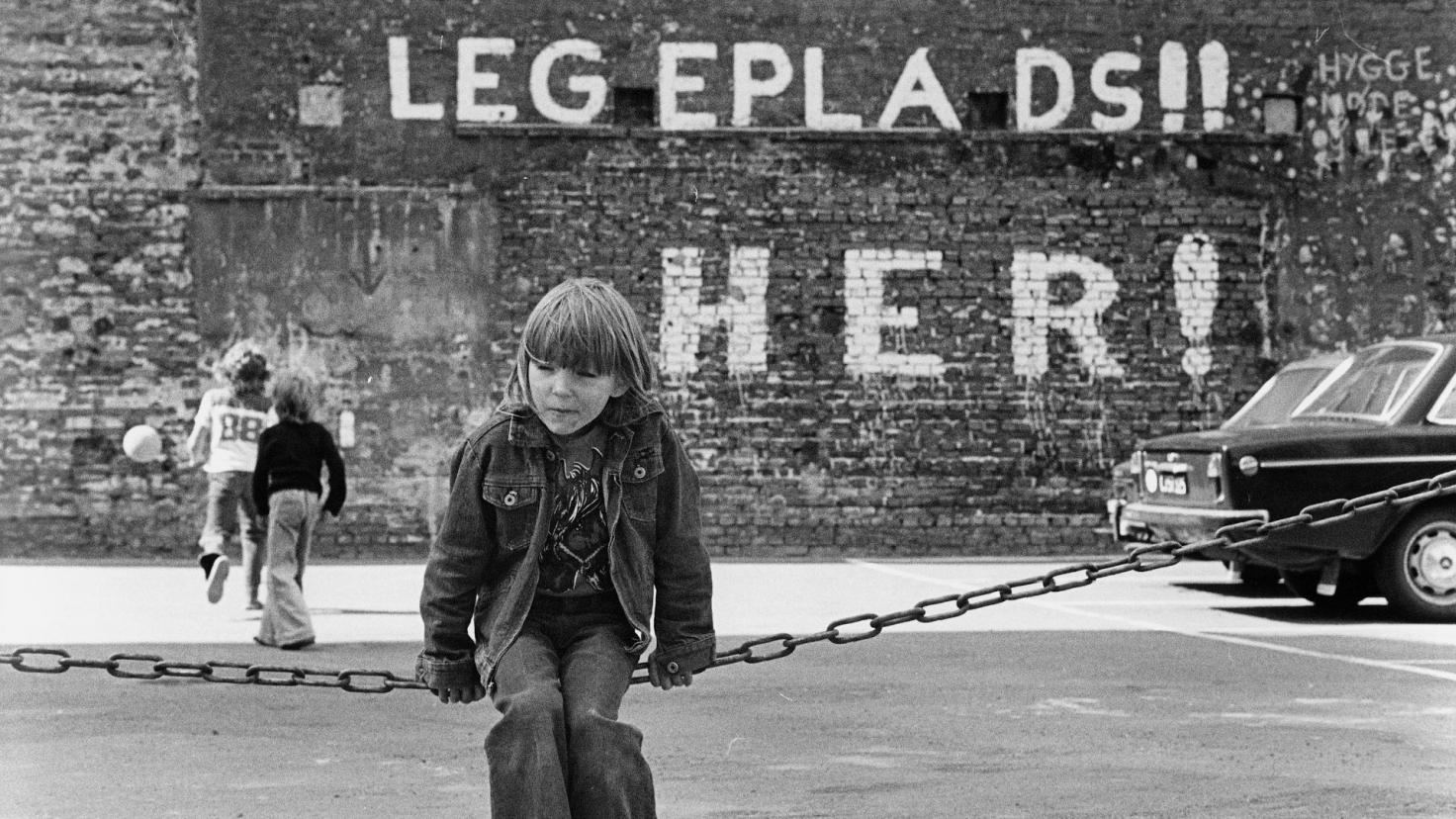 Sort/hvid foto af barn, foran et murmaleri med teksten Legeplads Her!